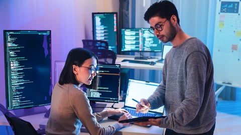 A man and a women working toghether with computer monitors with code in the background