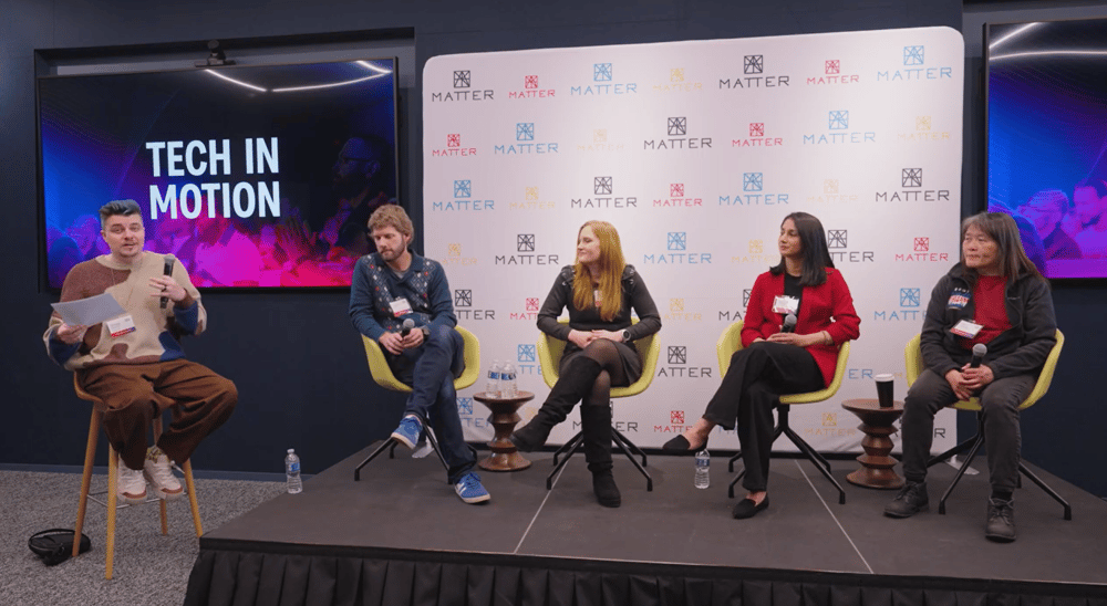 A panel discussion with five individuals, four seated on a stage and one speaking from a tall stool, under a backdrop with 