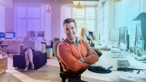 Person smiling at a desk in a modern, well-lit office with other workers in the background.